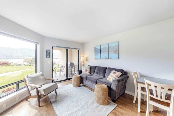 A modern living room with a grey sofa, white armchairs, and a dining table. There is a large window offering a view of a green landscape. - Bright 1BR Lakefront - Patio - Pool (Chelan)