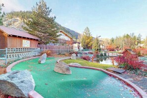 Um campo de mini golfe com superfície verde para tacadas, vários obstáculos e um edifício de madeira ao fundo.