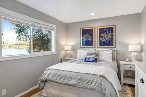 A bedroom with a large window, a bed with a striped bedspread, two bedside tables with lamps, and framed artwork on the wall. - Large 2BR Lakefront - Hot Tub - Dock - Patio (Chelan)
