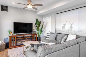 A living room with a grey sofa, a flat-screen TV, a ceiling fan, a fireplace, and a large framed picture of deer.