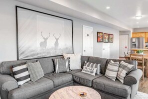 A modern living room with a sectional sofa, a coffee table, and a large framed picture of deer. - Large 2BR Lakefront - Hot Tub - Dock - Patio (Chelan)
