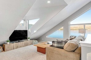 A modern loft bedroom with a large skylight, a flat-screen TV, a sofa, and a view of the outdoors. - Large 2BR Lakefront - Hot Tub - Dock - Patio (Chelan)