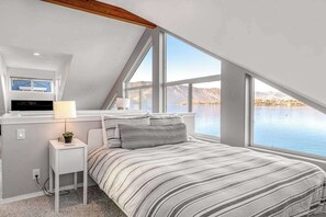 A bedroom with a bed, a nightstand, a lamp, and a window overlooking a body of water.