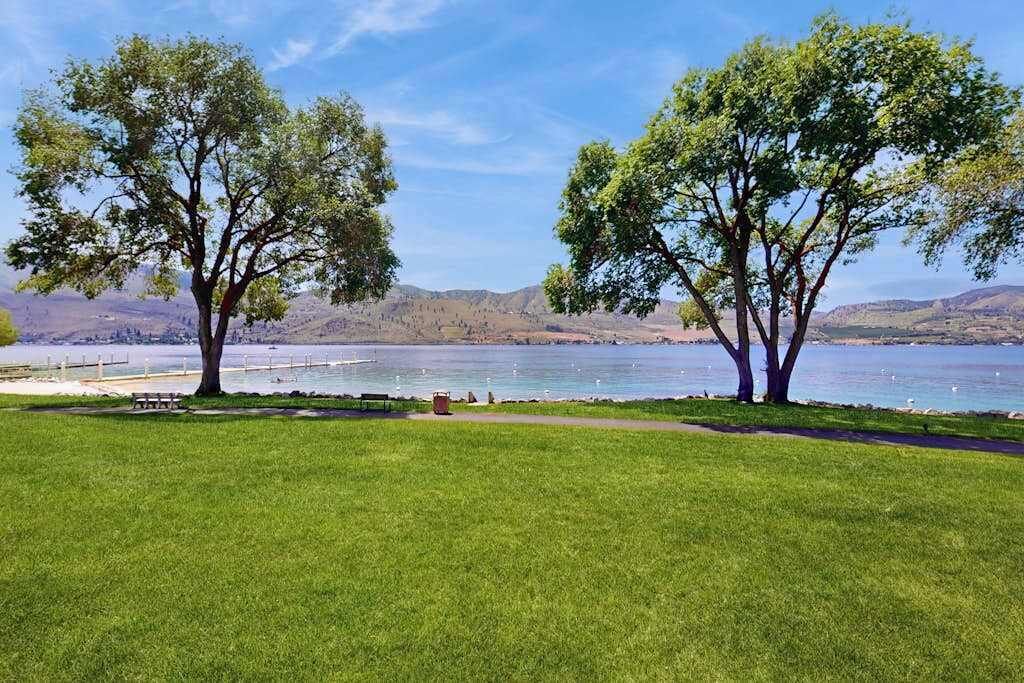 Un parc avec deux grands arbres, un banc et une vue sur un lac, avec des montagnes en arrière-plan.