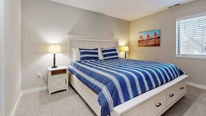 Een slaapkamer met een bed, nachtlampen, een nachtkastje, een schilderij en een raam met jaloezieën.