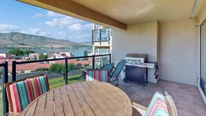 Een balkon met uitzicht op water en heuvels, een barbecue en een tafel met stoelen.