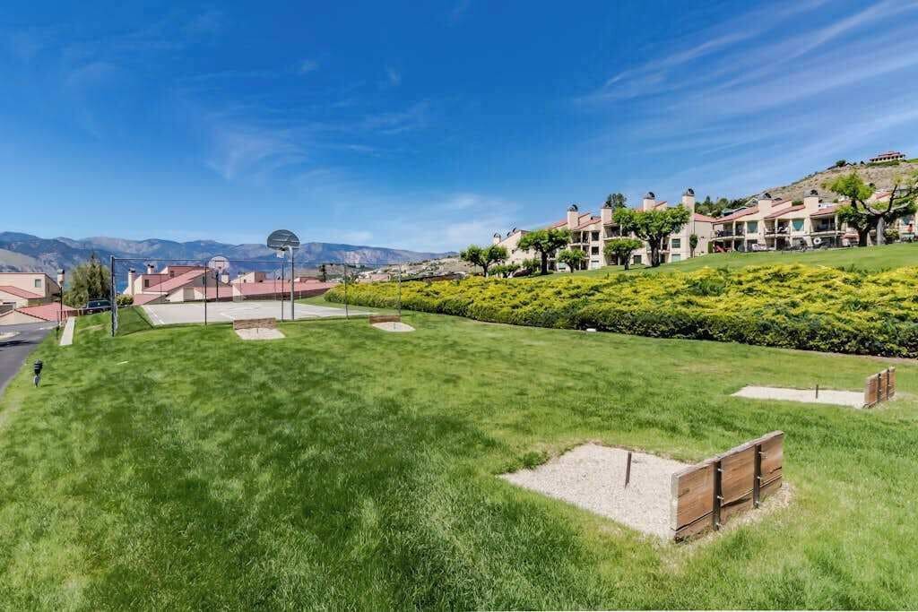 A well-maintained grassy area with a basketball court, a wooden bench, and a view of a residential area with houses and a hilly landscape in the background.