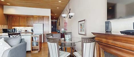 A modern kitchen with wooden cabinets, a dining area with a glass table and striped chairs, and a built-in fireplace.