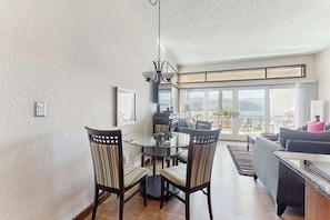 A dining area with a glass table and chairs, a kitchen in the background, and a balcony with a view. - Stylish 1BR Lakefront - Patio - Pool (Chelan)