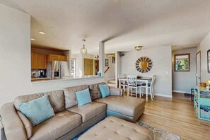 Ein modernes Wohnzimmer mit einem beigefarbenen Ecksofa, einem Essbereich mit weißem Tisch und Stühlen sowie einer Küche mit Holzschränken und Edelstahlgeräten.