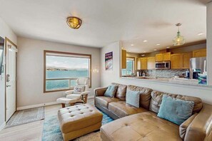 A modern living room with a large window, a sofa, a coffee table, and a kitchen area with wooden cabinets and a view of the sea. - Huge 3BR Lakefront - Hot Tub - Dock - Deck (Chelan)