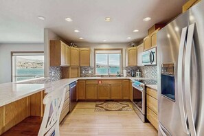 A modern kitchen with wooden cabinets, a stainless steel refrigerator, and a large window overlooking a scenic view. - Huge 3BR Lakefront - Hot Tub - Dock - Deck (Chelan)