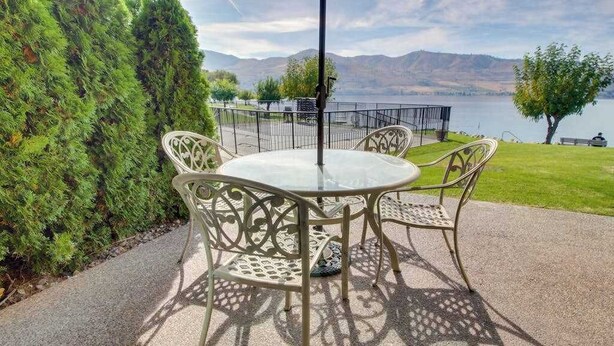 Eine Terrasse mit rundem Tisch und Stühlen, einem Metallzaun und Blick auf einen See mit Bergen im Hintergrund.