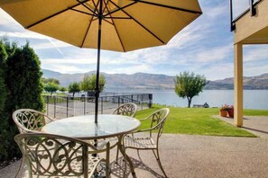 Un patio con una mesa, sillas, una sombrilla y vistas a un lago y montañas.
