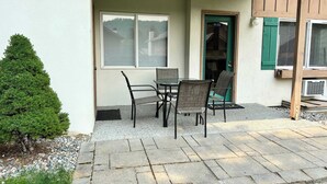 Een terras buiten met een tafel en stoelen, een deur met een raam, en een kleine boom.