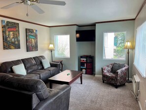 Ein Wohnzimmer mit einer Couch, einem Sessel, einem Couchtisch und einem Fernseher. Zwei Fenster mit Jalousien, ein Deckenventilator und Kunstwerke an den Wänden sind ebenfalls vorhanden.