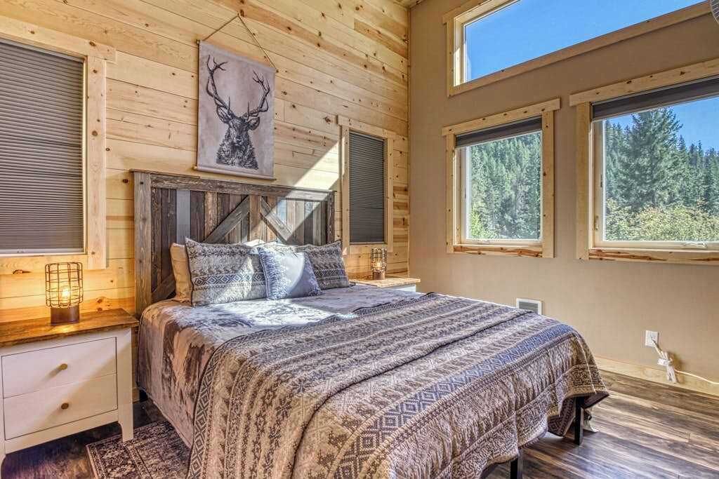 A bedroom with a bed, a wooden headboard, a wall-mounted deer artwork, and large windows with a forest view.