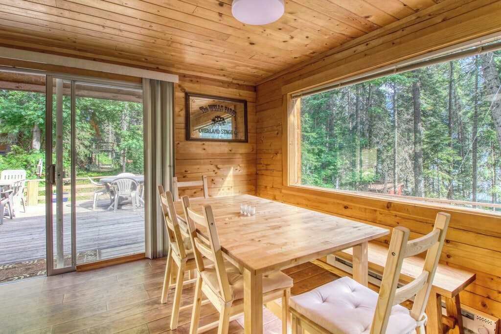 A wooden interior with a dining table and chairs, a large window with a forest view, and a door leading to an outdoor deck.