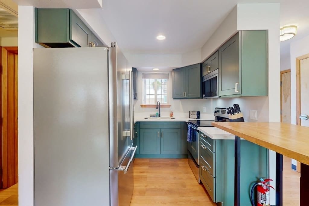 A modern kitchen with a stainless steel refrigerator, teal cabinetry, and a wooden countertop.