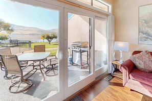 A room with a sliding glass door leading to an outdoor patio with a table and chairs, a grill, and a couch with a lamp.
