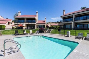 Ein Swimmingpool mit einer Metallleiter, umgeben von Liegestühlen und Sonnenschirmen, mit Gebäuden im Hintergrund.