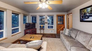 A living room with a ceiling fan, a television, a sofa, a coffee table, and large windows. - Woodland 2BR Mountainview Dog Friendly - Deck (Leavenworth)