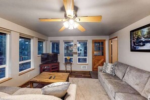 A living room with a ceiling fan, a television, a sofa, a coffee table, and large windows. - Woodland 2BR Mountainview Dog Friendly - Deck (Leavenworth)