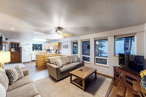 A living room with a sofa, coffee table, TV stand, and a kitchen area in the background. - Woodland 2BR Mountainview Dog Friendly - Deck (Leavenworth)