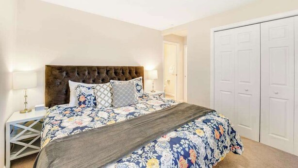 Quarto com cabeceira estofada, colcha florida, mesa de cabeceira com abajur e guarda-roupa branco.