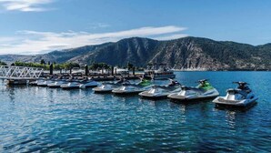 Marina com vários barcos pequenos atracados, um cais de madeira e montanhas ao fundo.