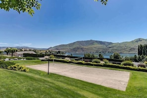 A tennis court with a net, surrounded by well-maintained grass and shrubs, with a scenic view of hills and a lake in the background. - 2BR Lakefront Condo - Balcony - Pool - Firepit (Chelan)
