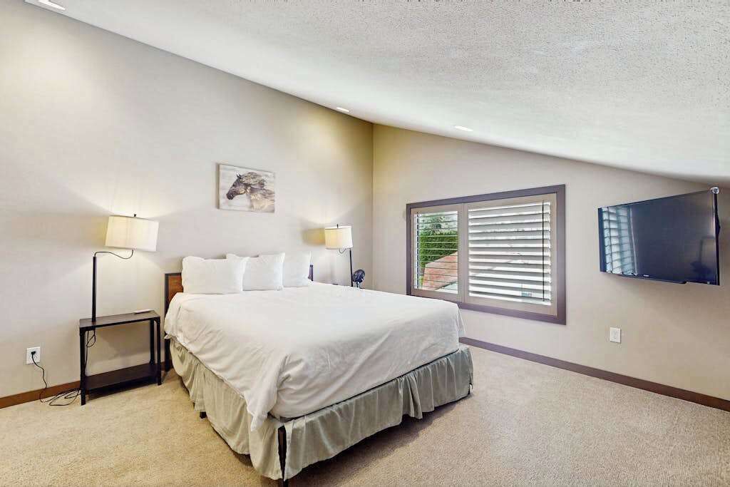 A bedroom with a bed, bedside lamps, a mounted TV, and a window with blinds.