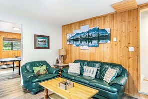 A cozy living room with a green leather sofa, a wooden coffee table, and a wall-mounted painting of a mountain landscape. - Alpine 3BR with fenced yard, hot tub, & Ping-Pong (Leavenworth)
