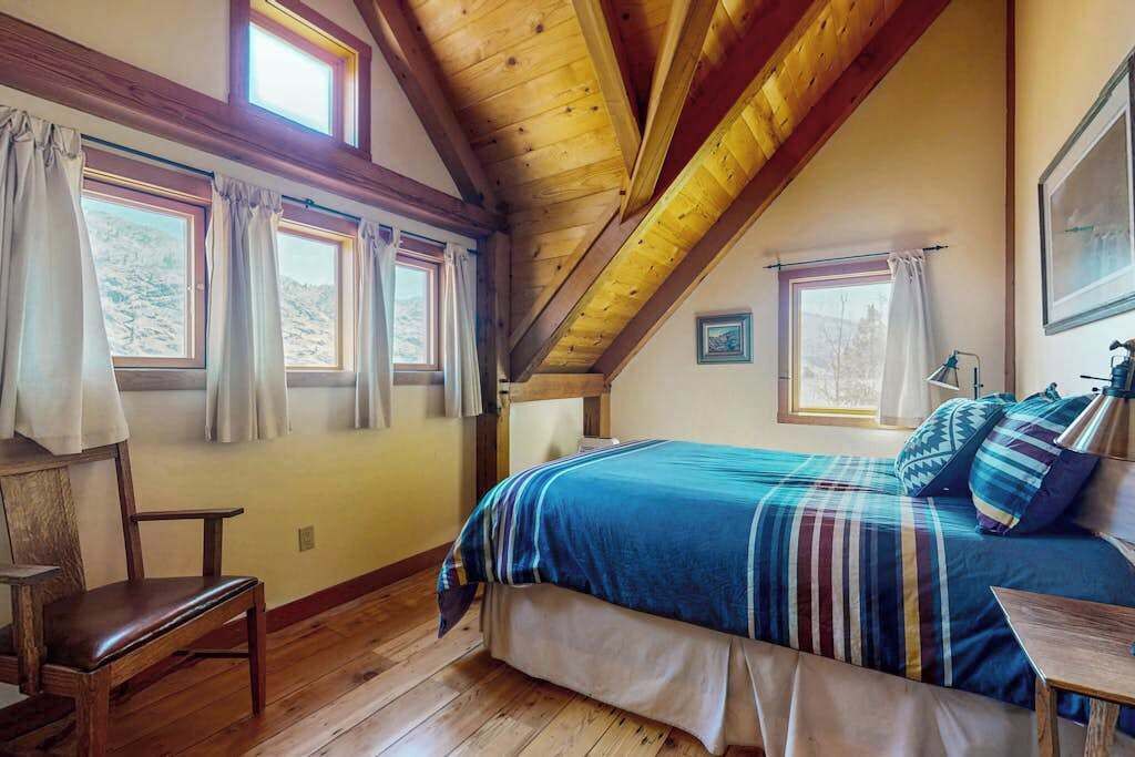 Ein Schlafzimmer mit Holzboden, einem Bett mit gestreifter Decke, einem Sessel und zwei mit Vorhängen versehenen Fenstern.