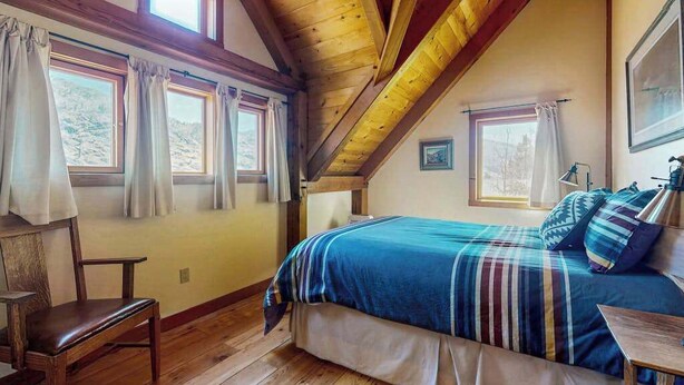 A bedroom with a wooden ceiling, a bed with a striped blanket, a chair, and two windows with curtains. - Unique 2BR Lakefront - Hot Tub - Dock - Deck (Chelan)