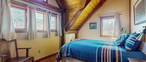 Un dormitorio con techo de madera, una cama con manta a rayas, una silla y dos ventanas con cortinas.