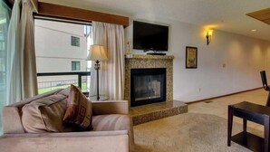 A living room with a sofa, a television mounted above a fireplace, and a view of a building outside. - 2BR Lakefront - Pool - Tennis - Hot Tub - Balcony (Chelan)
