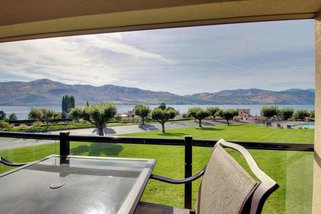 A balcony with a view of a lake and mountains, a table, and chairs.