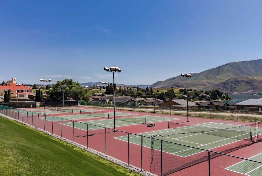 Ein Tennisplatz mit mehreren Spielfeldern, umzäunt, mit Häusern und Bergen im Hintergrund.