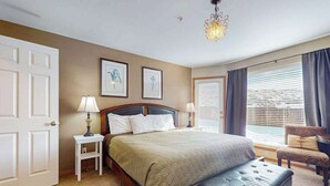 Ein Schlafzimmer mit einem Bett, Nachttischen, einer Bank, einer Chandelüster und einem Fenster mit Vorhängen.