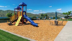 Ein Spielplatz mit Rutsche und Klettergerüsten, umgeben von einem mit Kies bedeckten Bereich und Blick auf die Hügel.