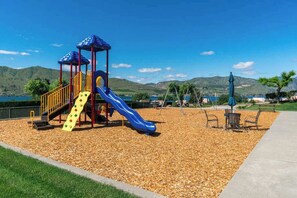 Ein Spielplatz mit Rutsche und Klettergerüsten, umgeben von einem mit Kies bedeckten Bereich und Blick auf die Hügel.