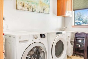 A laundry area with two white washing machines, a wooden cabinet, and a window with a view of trees.