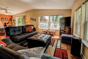 A living room with a sofa, ottoman, coffee table, TV stand, and a view of the outdoors.