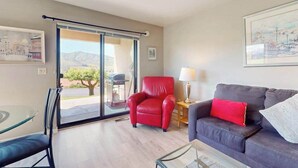Ein Zimmer mit Glastür zu einem Außenbereich, ein roter Sessel, eine dunkelgraue Couch mit einem roten Kissen und ein Tisch mit einer Lampe.