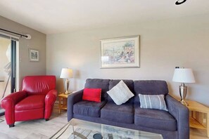 A living room with a red chair, a dark sofa with pillows, a glass coffee table, and two lamps on side tables. - Luxe 1BR Lakeview - Patio - Pool - (Chelan)