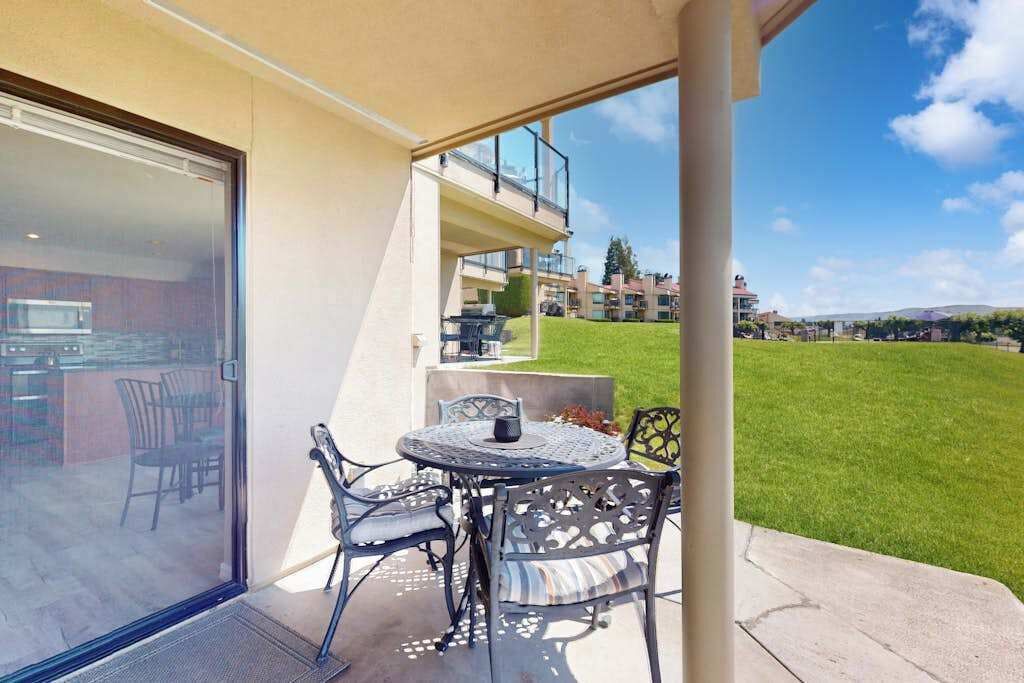 A patio with a table and chairs, a glass door leading to a kitchen, and a view of a green lawn and buildings in the background.