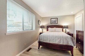 A bedroom with a bed, two bedside tables, a nightstand, a window with blinds, and a door. - Luxe 1BR Lakeview - Patio - Pool - (Chelan)
