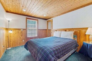 Une chambre avec un plafond en bois, un lit recouvert d’une couverture à carreaux, une table de chevet avec une lampe et une fenêtre avec des stores.