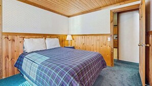 Ein Schlafzimmer mit Holzbalkendecke, einem Bett mit karierter Bettdecke, einem Nachttisch mit Lampe und einer Tür zu einem anderen Raum.